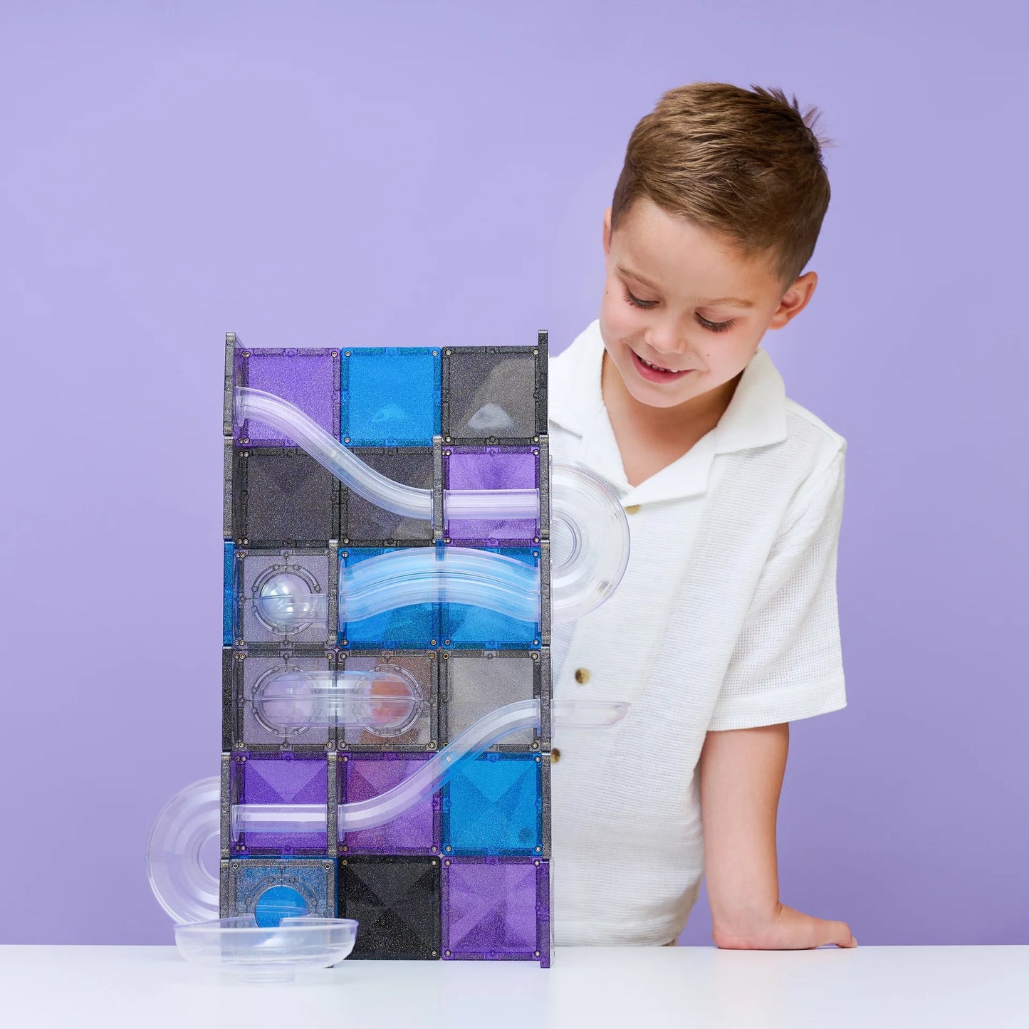 A young boy in a white polo shirt smiles while looking at a Connetix Ball Run Galaxy magnetic marble run toy built from purple, blue, and black translucent tiles on a white table against a solid lavender background.
