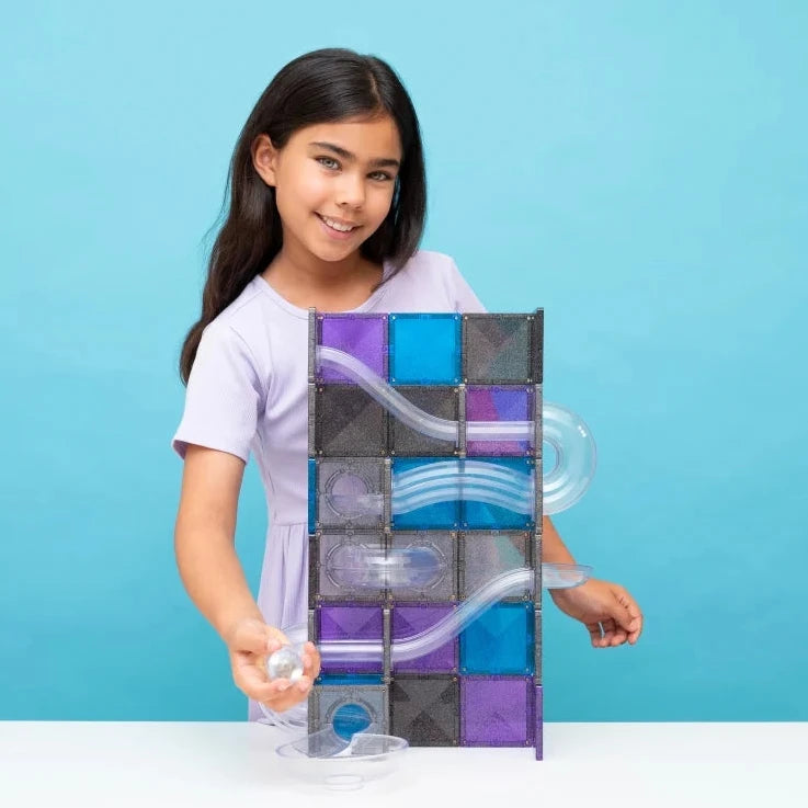 A young girl with long dark hair, wearing a light purple t-shirt, stands behind a Connetix magnetic tile Galaxy Pack track toy, holding a small clear marble. The toy consists of black and purple magnetic tiles with clear plastic tracks.